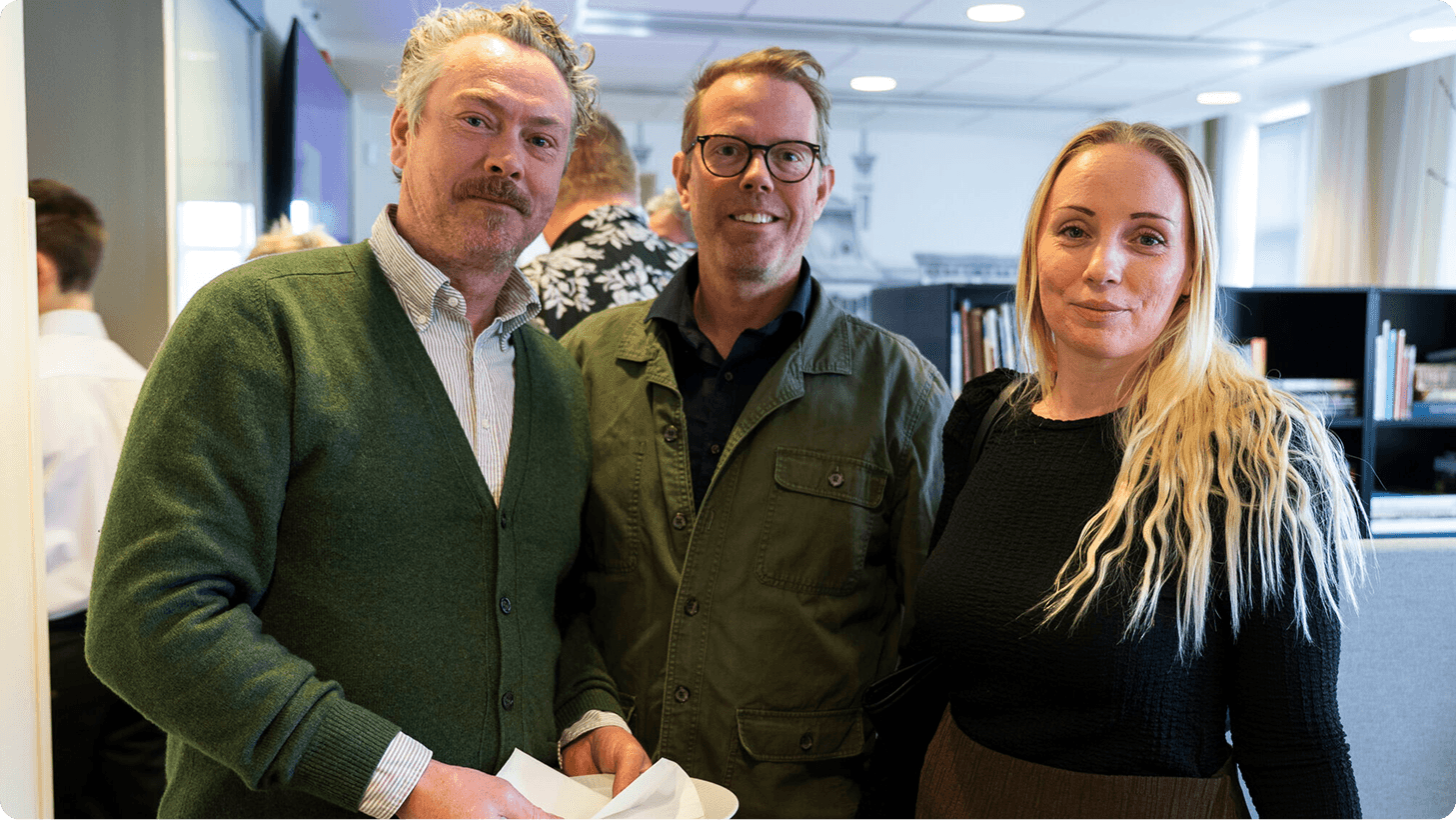Three people, including a man with a mustache and a blonde woman, pose together.