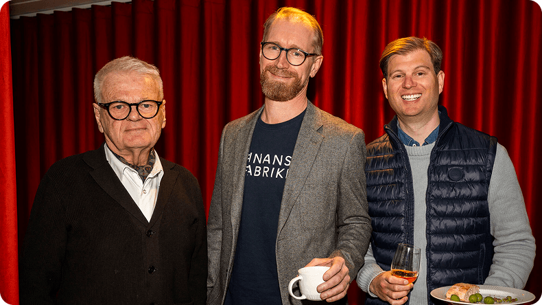 Three smiling men, dressed in casual event attire, stand in front of a red curtain.